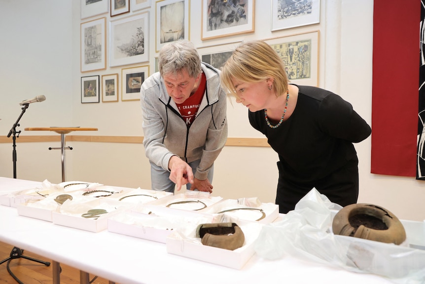 A man and a woman lean over a table to inspect an array of ancient jewellery. 