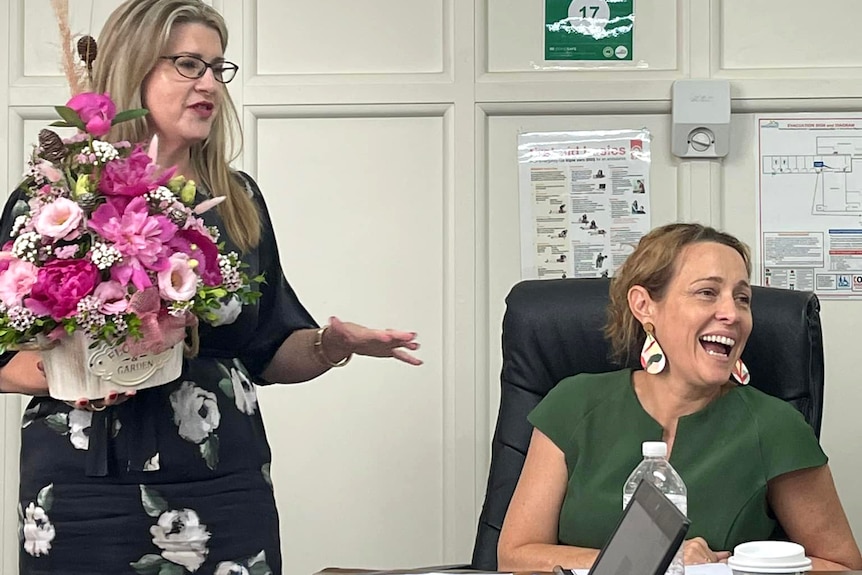 two women in an office, both have blonde hair, one is standing and holds a bunch of flowers, the other is sitting and laughing