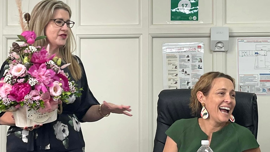 two women in an office, both have blonde hair, one is standing and holds a bunch of flowers, the other is sitting and laughing