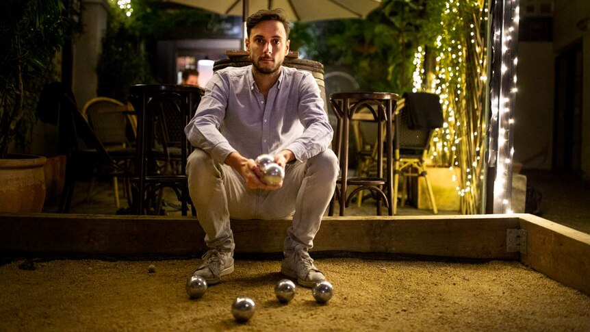 A man sits on a small wall with small metal balls on the ground and in his hands.