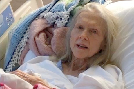 An elderly woman lays on Queensland Health linen, looking frail, grey hair.