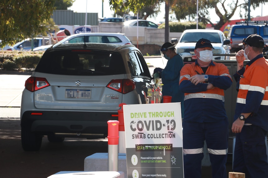 Two workers in hi-vis clothing stand at a drive-through COVID testing clinic in Joondalup with a car behind them.