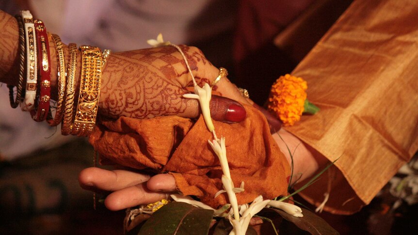 A woman's hand, covered in henna and her arm adorned with bangles, rests on top of a man's hand.