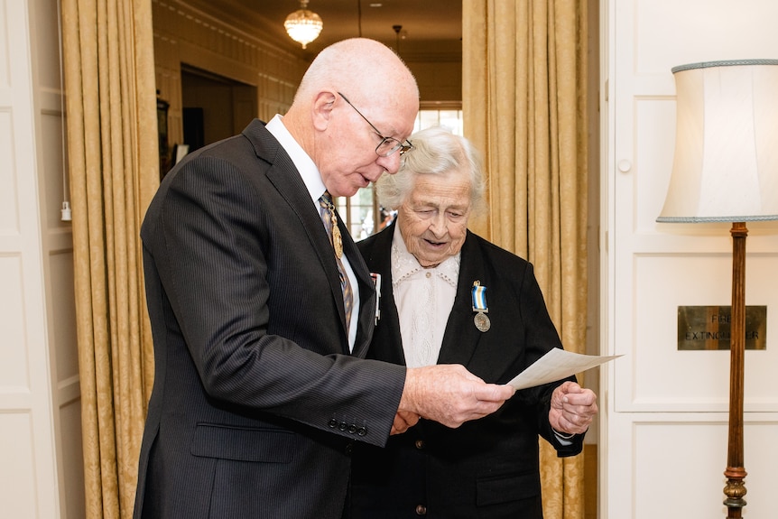 An elderly woman with a medal on her chest looks at a piece of paper with the Governor-General 
