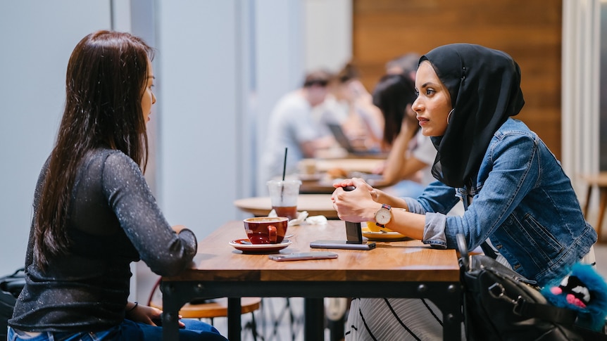 Two women sitting at a table drinking coffee and talking. 