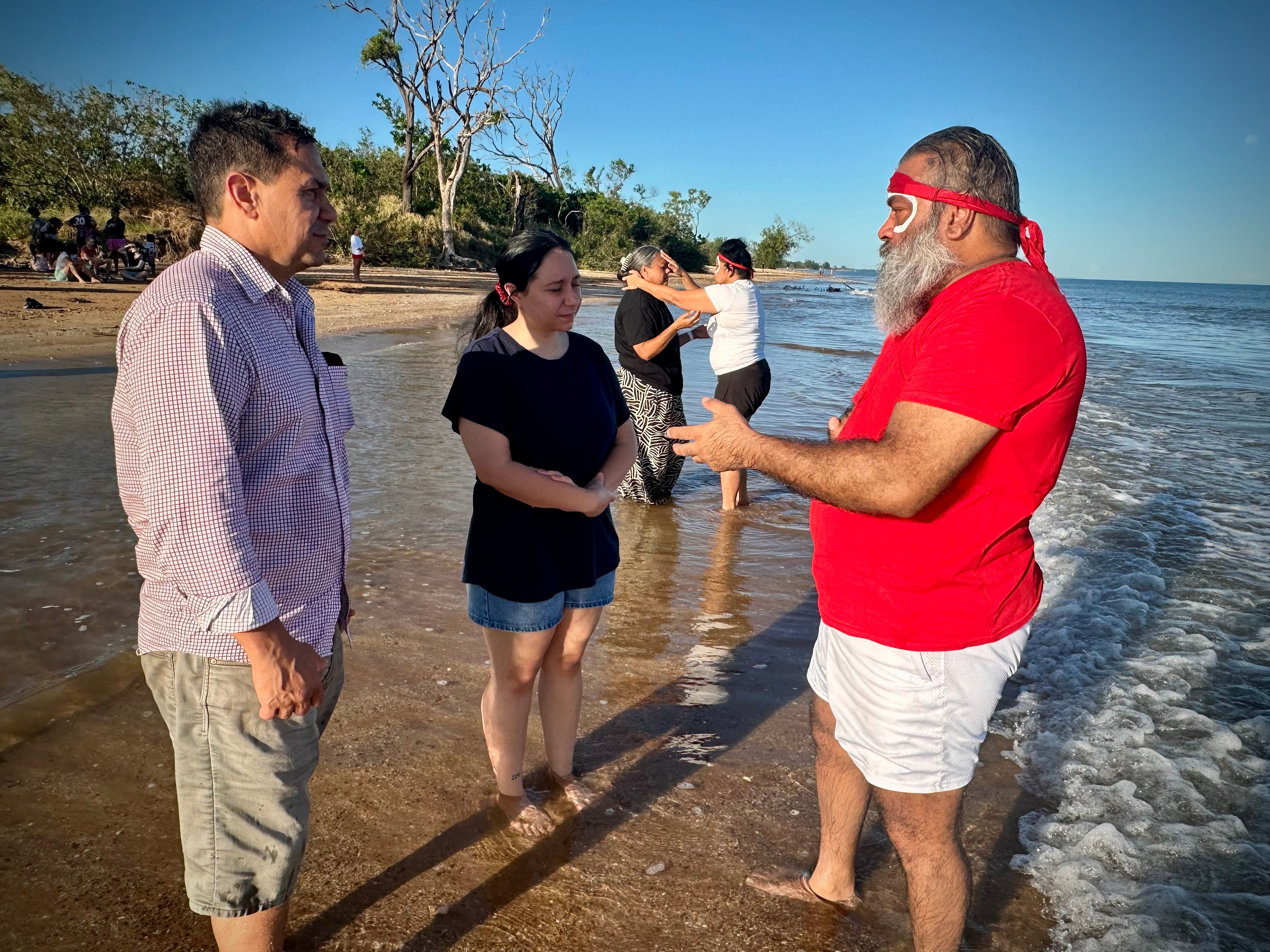 A traditional owner dressed in red, speaking to people as they stand on a beach near the edge of the ocean.