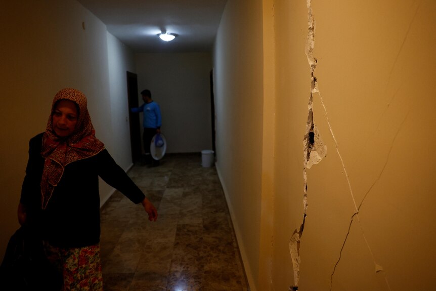 A woman walks past a wall that has a large crack in it. 