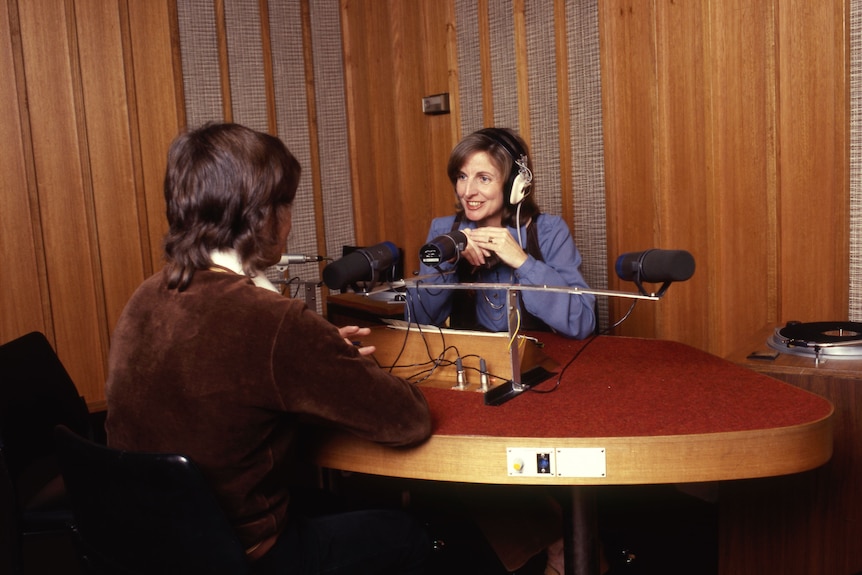 A colour image from 1979 of Caroline Jones seated at a desk wearing headphones with her male inteview subject facing away 