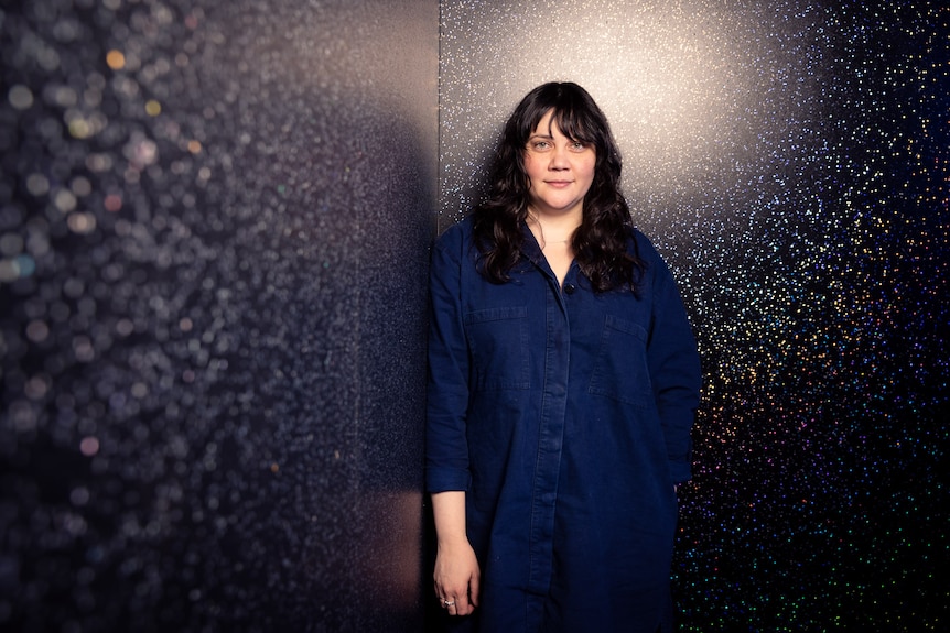 Shari Sebbens stands in the corner of a glittery black theatre set, facing the camera , with a slight smile