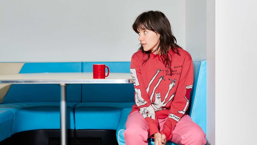 Courtney Barnett wearing a red top and pink trousers sitting on a blue bench seat cafe table.