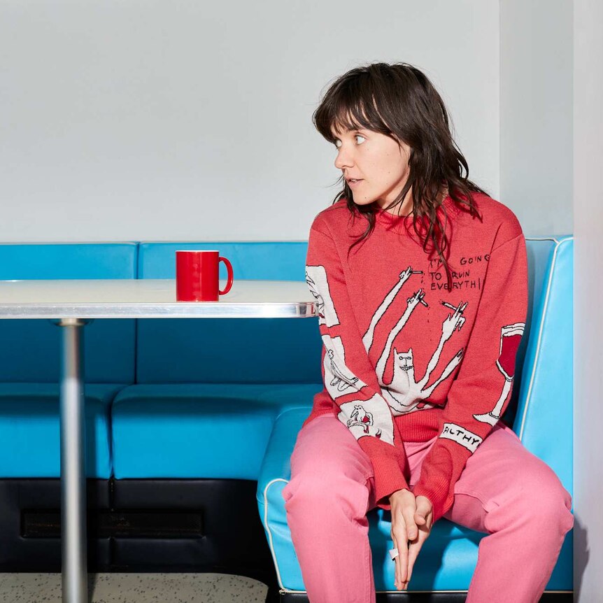 Courtney Barnett wearing a red top and pink trousers sitting on a blue bench seat cafe table.