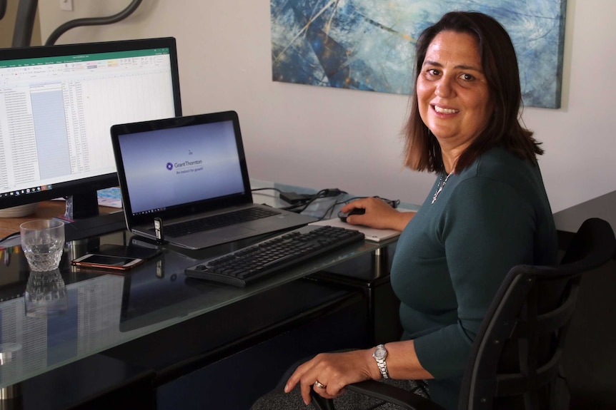 Ms Eagan sits in a desk chair in front of her computer, a spreadsheet in front of her.