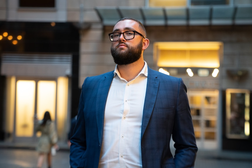 A  man with a beard and glasses poses in front of buildings on a city street