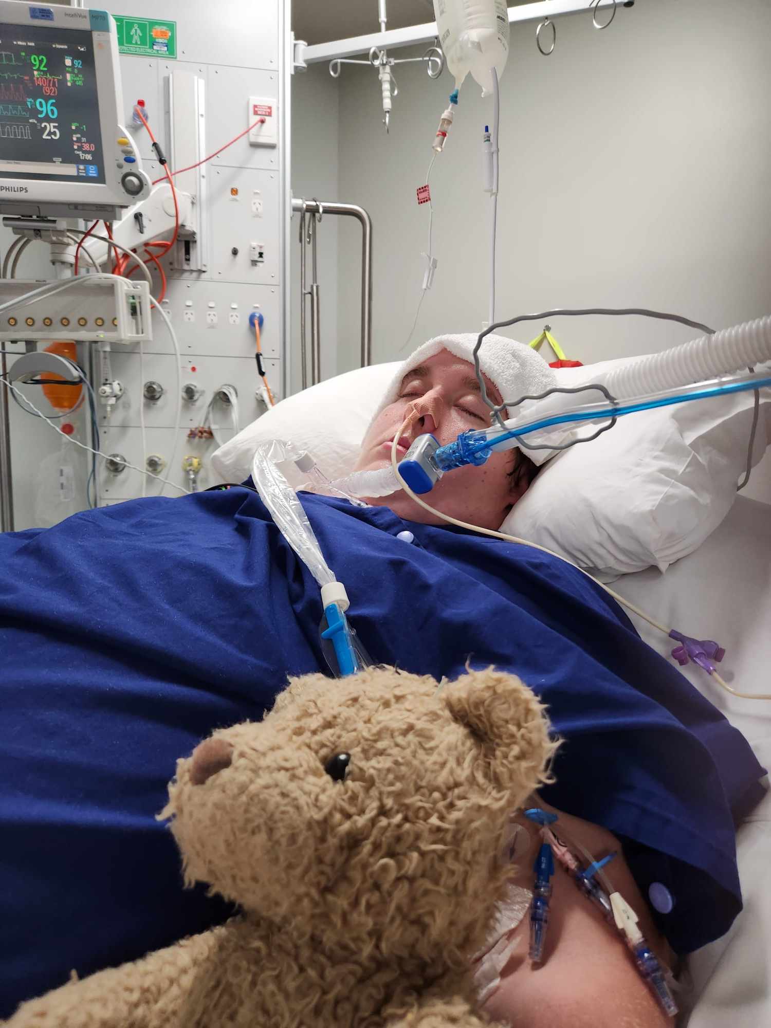 A young man in a hospital bed with tubes connected to his throat and teddy bear nearby