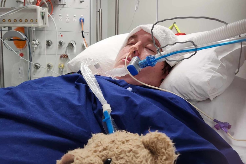 A young man in a hospital bed with tubes connected to his throat and teddy bear nearby