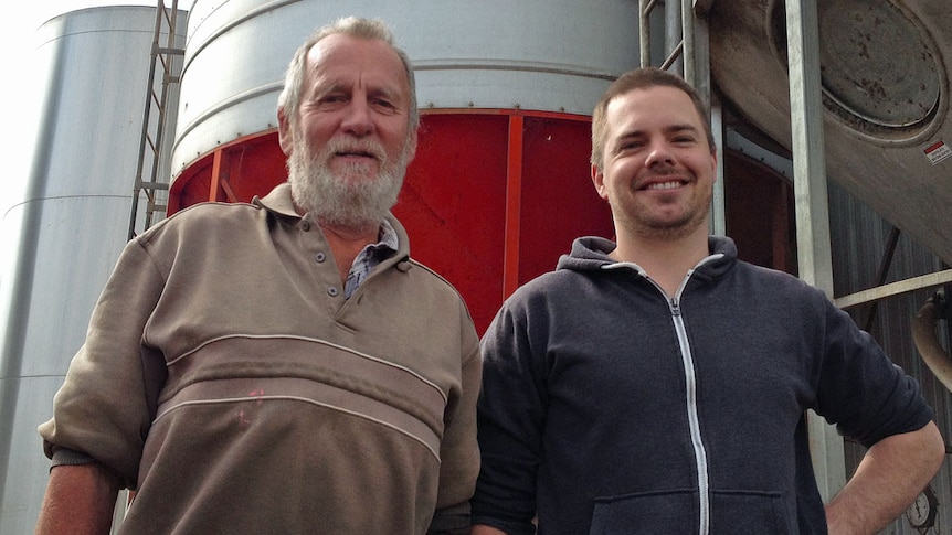 Father and son stand together in front of grain silos and their rotary dairy
