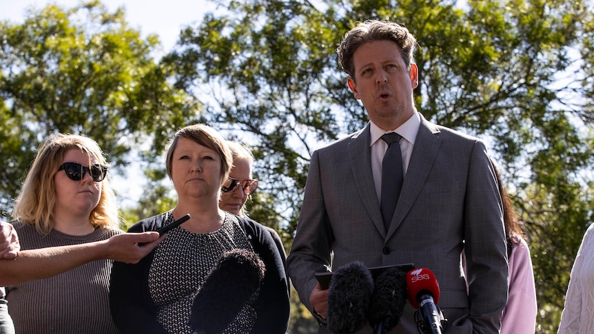 A man stands in front of microphones with several women standing next to him.