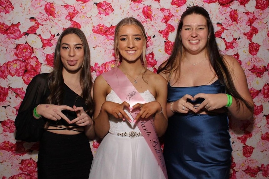 Jemma Borcich wears a white dress with a pink sash, she and two female friends smile and make love heart signs