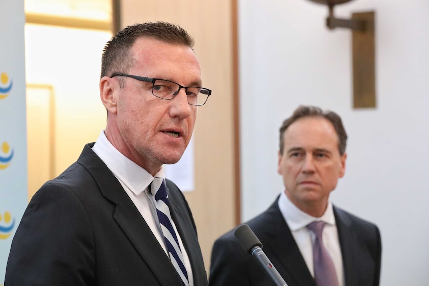 Greg Hunt watches on as Geoff Parker addresses the media. Mr Park is wearing glasses and a blue striped tie.