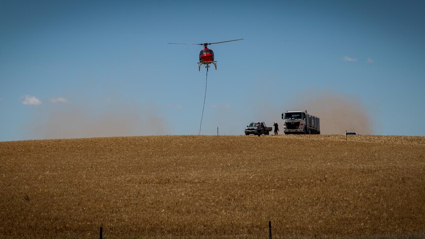 A red helicopter in the sky with a long rope attached with dust around it. Two vehicles are on the ground