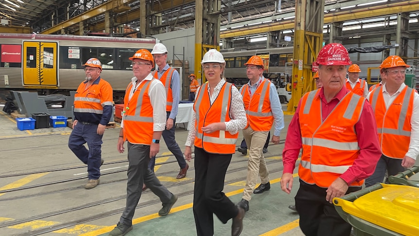 Premier wears orange vest and white hard hat. Surrounded by mean walking through a factory.