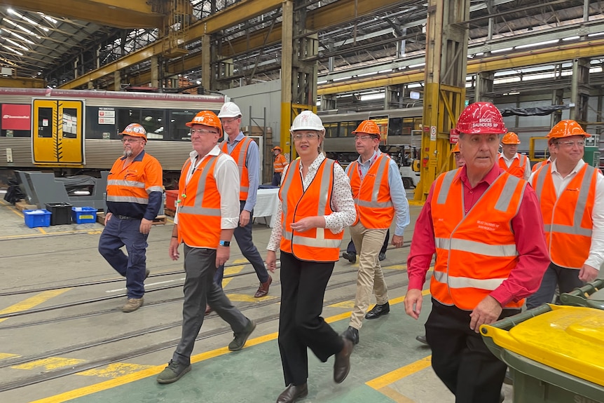 Premier wears orange vest and white hard hat. Surrounded by mean walking through a factory.