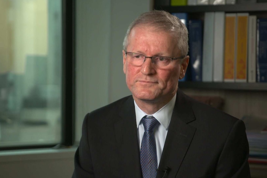 A man sitting in an office wearing glasses looks away from the camera with a neutral expression