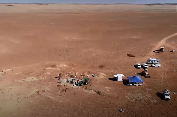 A drone's view of a dig: an aerial view of the desert expanse and a camp of vehicles, a large tent, machinery, people, and a dig