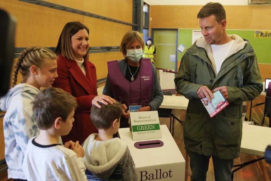 Three children and three adults stand next to a voting box in a polling booth.