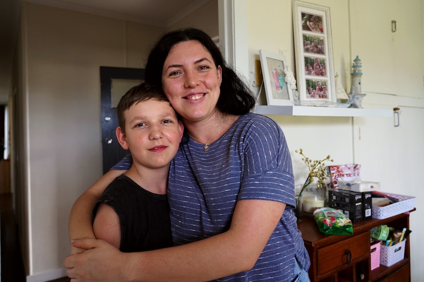 a woman smiles widely while embracing a young boy who also smiles, they're standing in a hall way