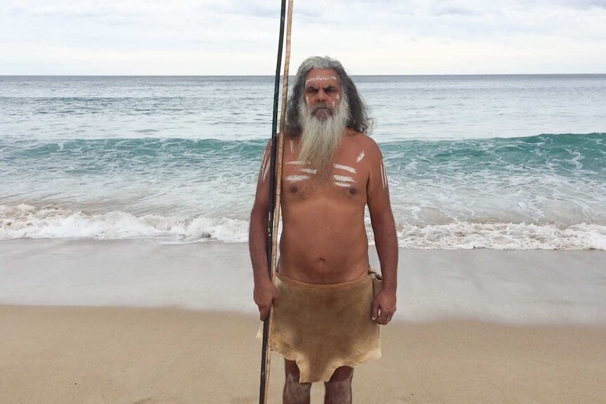 An Indigenous man stands wearing an animal skin on a beach.