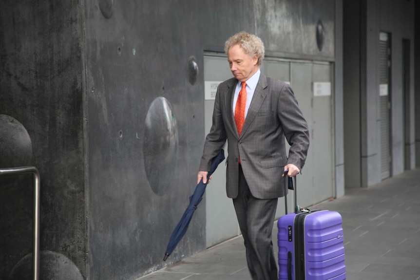 A man in a grey suit and an orange tie walks with a purple suitcase and a navy umbrella outside a building.
