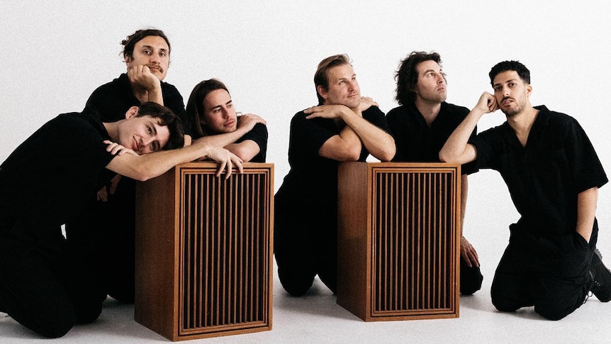 The six members of Mel Blue, all young men wearing black clothing, stand in front of a white background.