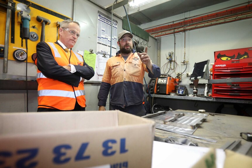 Wearing hi-vis vests and protective glasses, Bill Shorten talks with a factory worker who is pointing into the distance