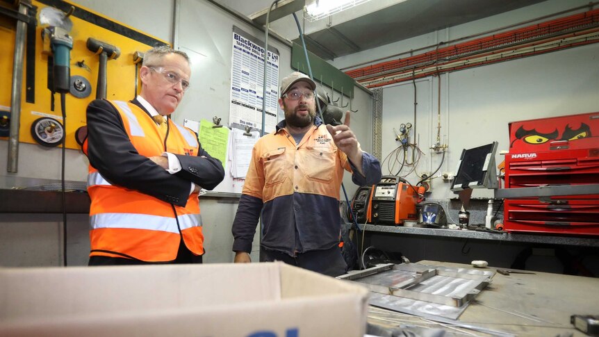 Wearing hi-vis vests and protective glasses, Bill Shorten talks with a factory worker who is pointing into the distance