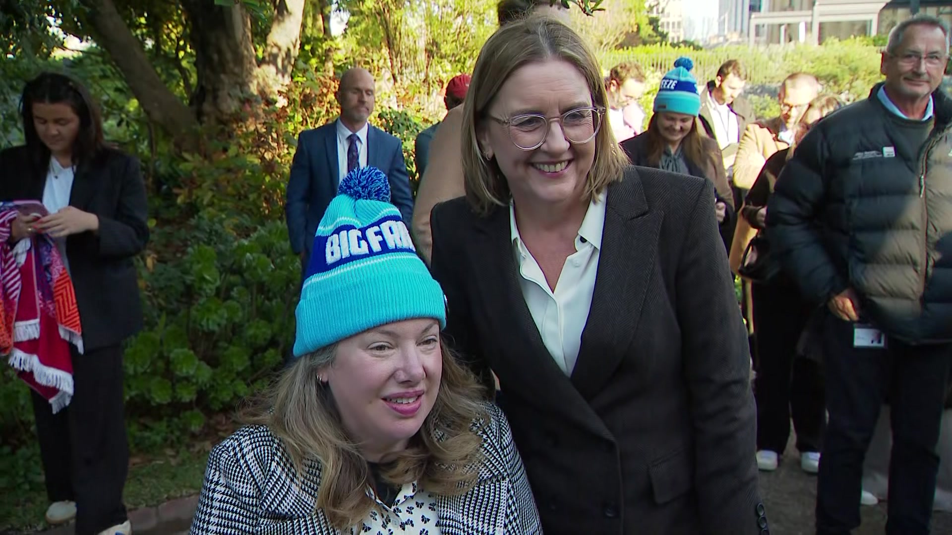 A woman in a blue beanie and checked blazer sits beside a woman with chin length light hair in a dark blazer who is standing.