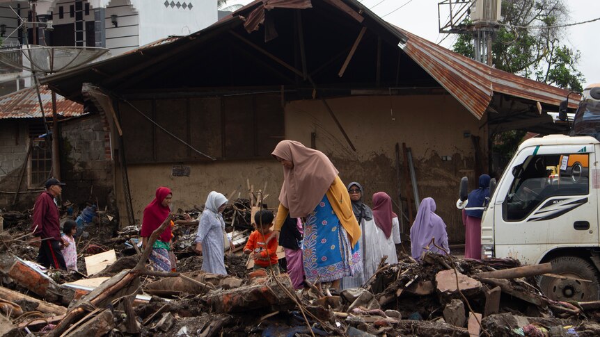 Group of people pick through rubble outside damaged buildings in a disaster zone.