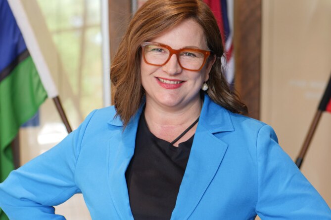 A woman wearing red-rimmed glasses and a blue jacket smiles at the camera