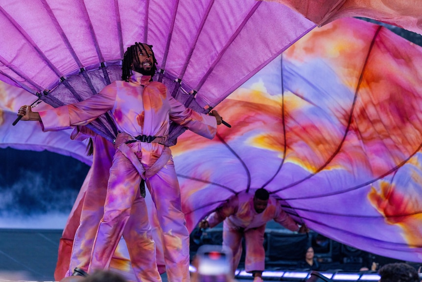 A back up dancer in a pink and purple jumpsuit with a large pink parachute behind him