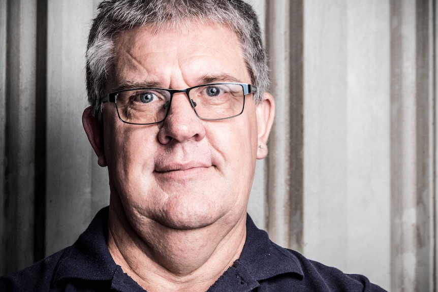 Allan Watson looks down the camera and stands in front of a metal shed wall.