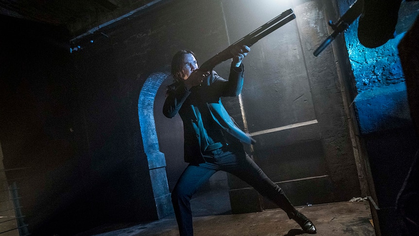 Colour wide angle still of Keanu Reeves dressed in suit and aiming raised gun in a dark space in John Wick: Chapter 3.