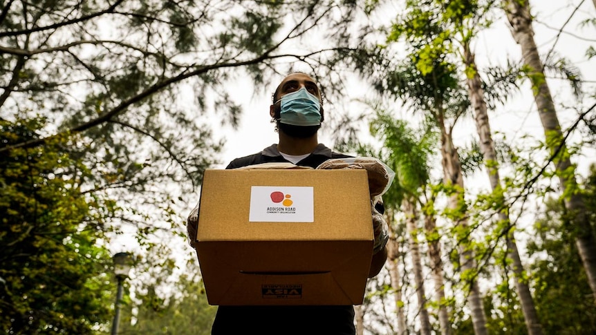 Man with face mask holding an Addison Road food hamper cardboard box walking down footpath with trees in background.