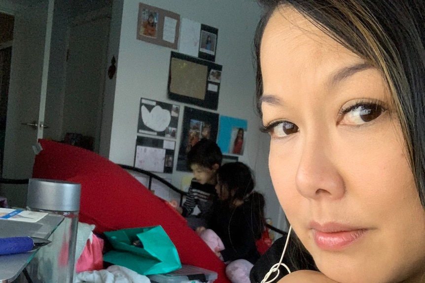 A woman looking at the camera, while her child works in the background.