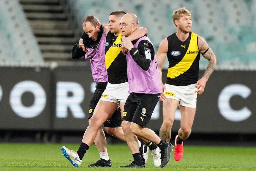 An AFL player is helped off the ground by two trainers after injuring his knee.