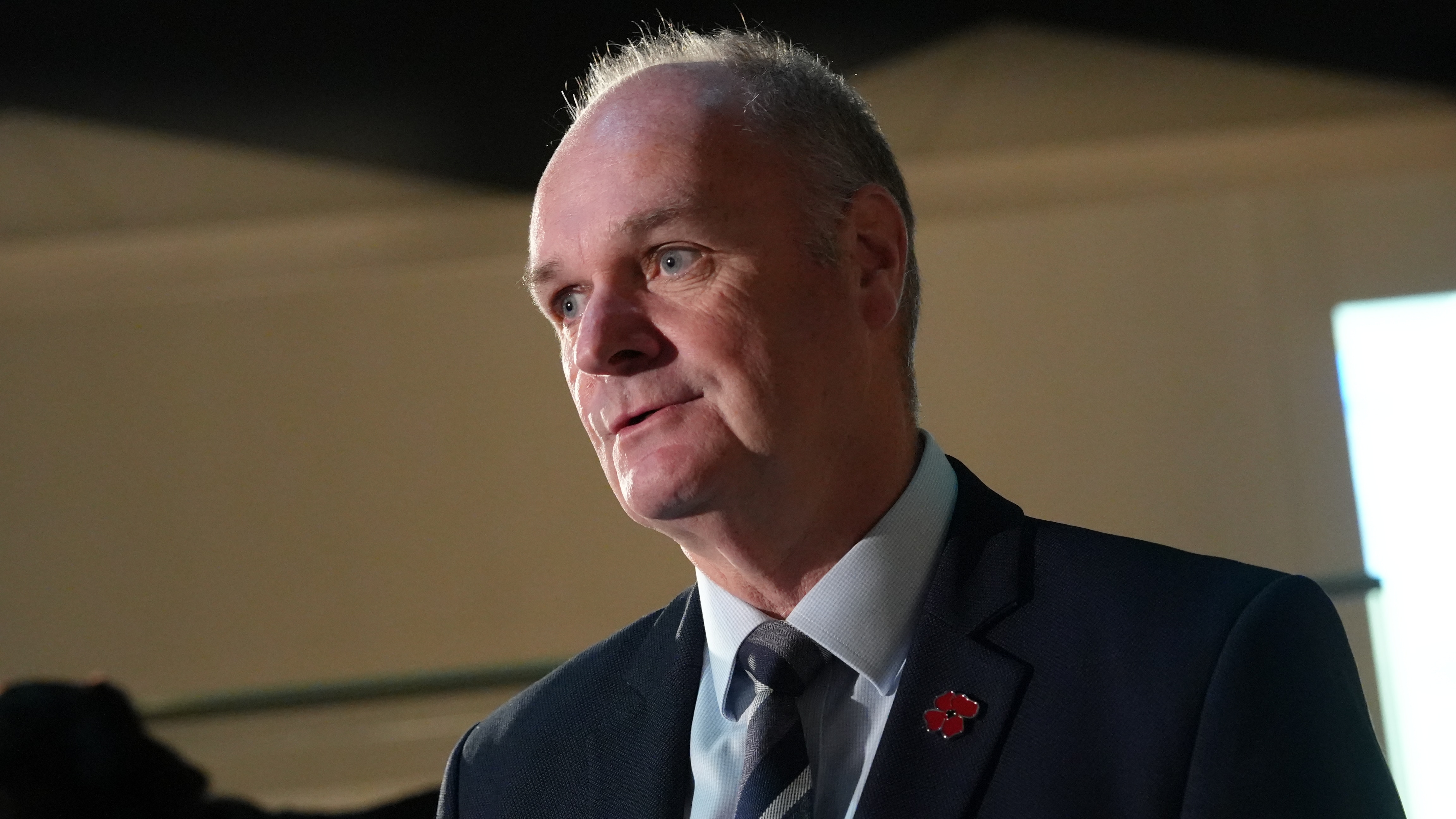 A man with greying haird, wearing a suit with a red poppy pin, speaks to people.