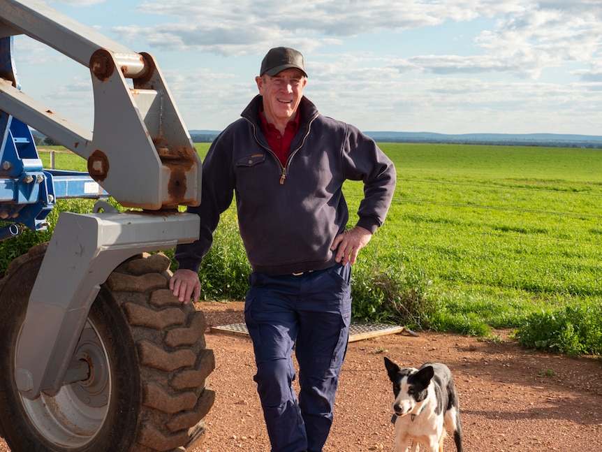 A farmer standing next to farm machinery in front of a green cropping paddock.