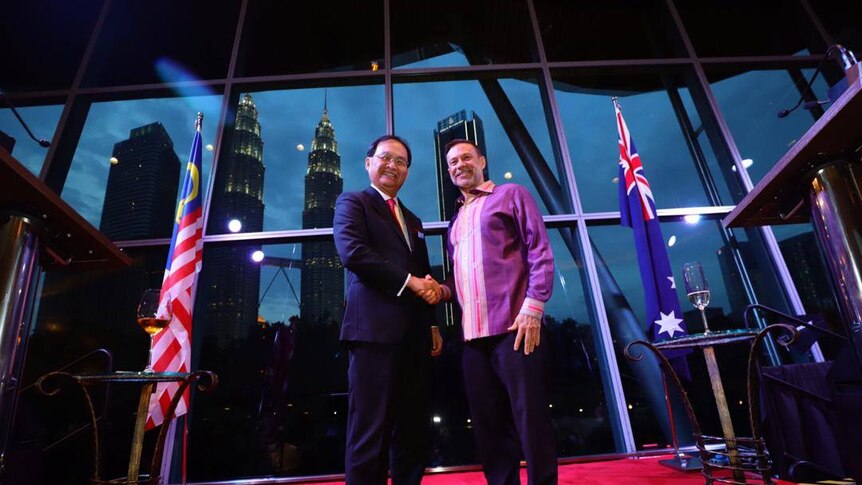 Two men stand on a stage in front of Malaysian and Australian flags and Kuala Lumpur's twin towers.