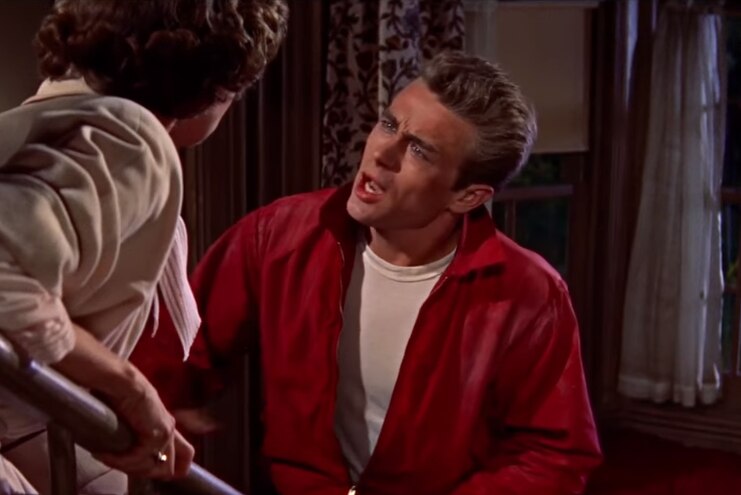 James Dean, who is wearing his red jacket, speaks to a woman on the stairs inside a house.