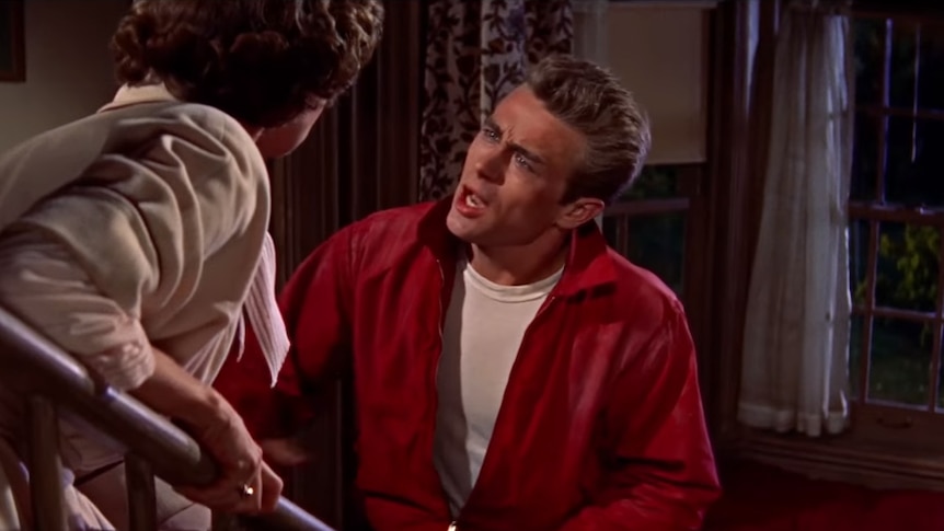 James Dean, who is wearing his red jacket, speaks to a woman on the stairs inside a house.