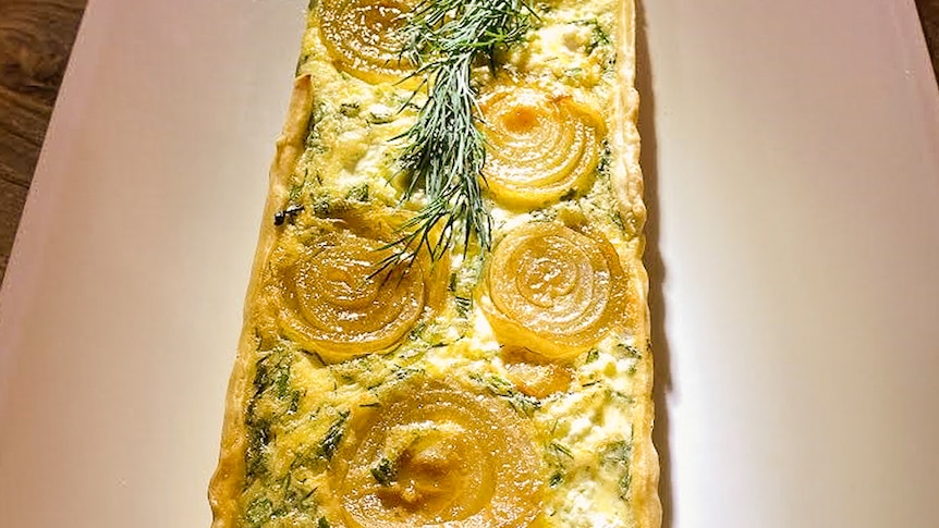 Image of a long, rectangular, shaped tart congtaining egg, onion and cheese displayed on a platter.